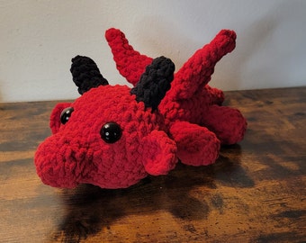May include: A red crocheted dragon plush toy with black horns and eyes. The dragon is lying on a wooden surface.