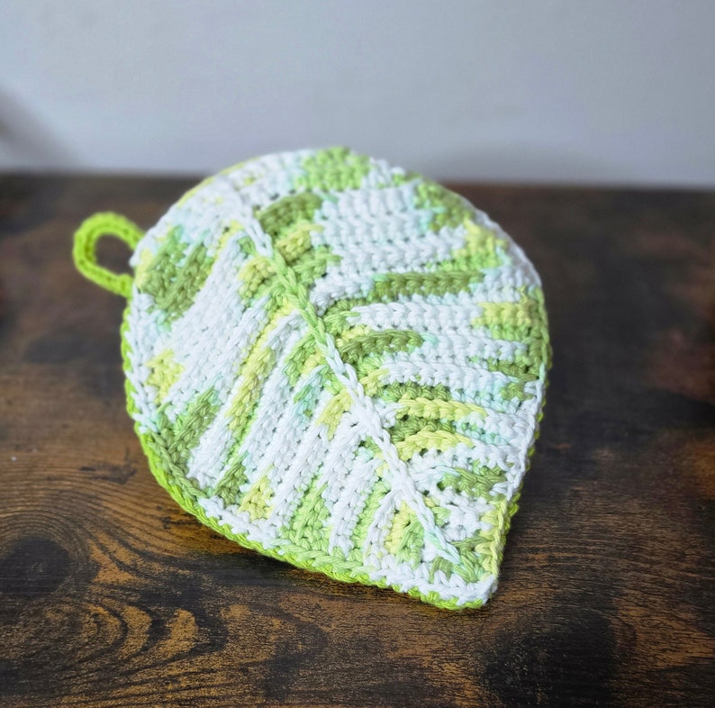 May include: A crocheted leaf-shaped pot holder in green and white. The pot holder has a loop for hanging.