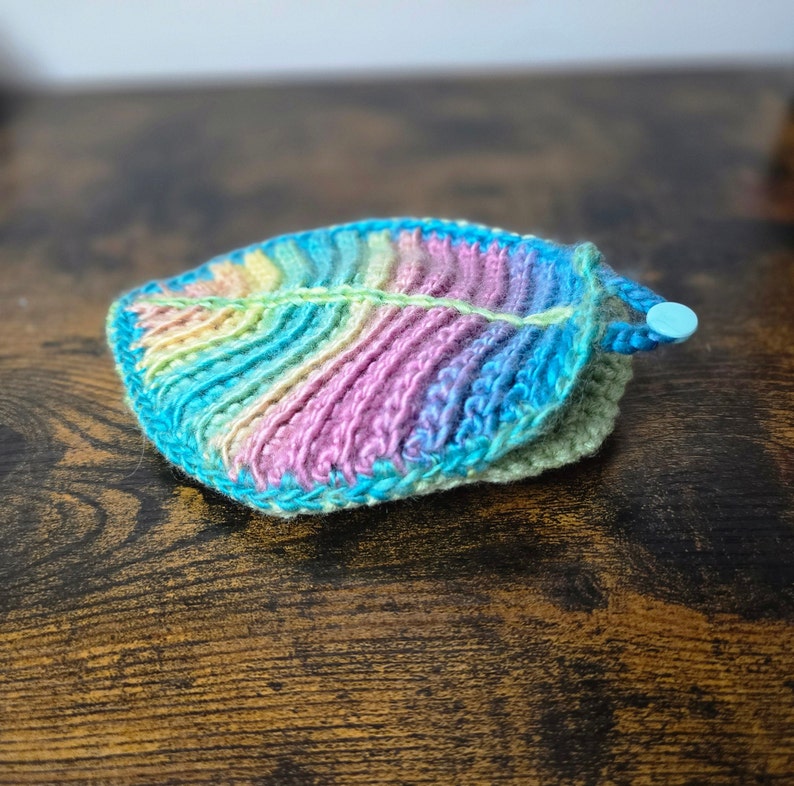 May include: A rainbow crocheted leaf with a blue border. The leaf is lying on a wooden surface.
