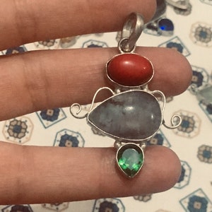 May include: A silver pendant featuring three gemstones. The top stone is oval and red, the middle stone is a large, irregular shape in shades of grey, and the bottom stone is a green teardrop shape.