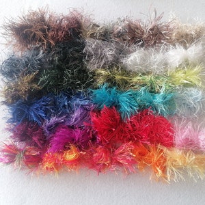 Puede incluir: Una colección de coloridos y esponjosos ovillos de lana en varios tonos, incluyendo rojo, rosa, naranja, azul, verde y morado. La lana tiene un aspecto texturizado y difuso, y está dispuesta sobre una superficie blanca.