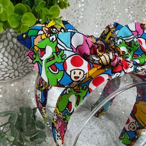 May include: A colorful headband featuring a bow with a vibrant print of video game characters. The headband has a clear plastic band and is decorated with images of Yoshi, Toad, and other characters on a blue background.