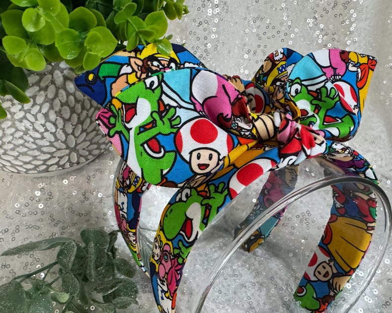 May include: A colorful headband featuring a bow with a vibrant print of video game characters. The headband has a clear plastic band and is decorated with images of Yoshi, Toad, and other characters on a blue background.