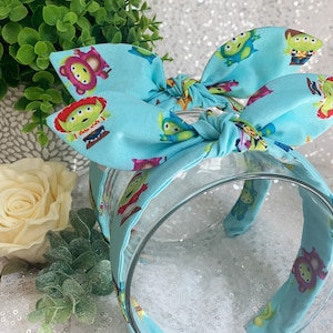 May include: A light blue headband with a bow, featuring a repeating pattern of green alien characters from the Toy Story franchise.