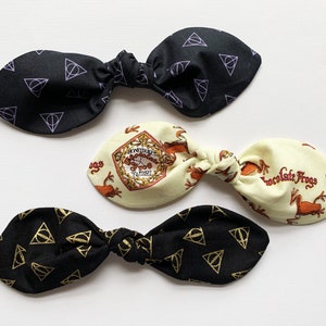 May include: Three fabric hair bows with different patterns. One bow is black with a purple triangle pattern, another is black with a gold triangle pattern, and the last is cream with a brown frog and clock pattern. The cream bow has the text "Chocolate Frogs" printed on it.