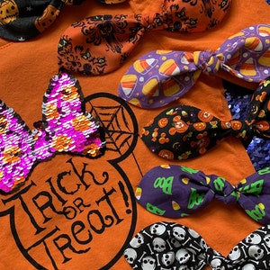 K&ouml;nnte beinhalten: Eine Sammlung von Halloween-Haarb&auml;ndern mit verschiedenen Mustern, darunter Candy Corn, K&uuml;rbisse, Totenk&ouml;pfe und ein Spukhaus. Die Schleifen sind auf einem orangefarbenen Shirt mit einem Mickey-Maus-Kopf und dem Text "Trick or Treat!" angeordnet.