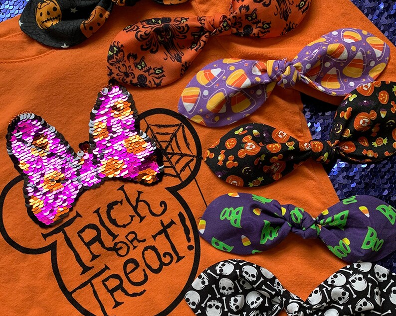 K&ouml;nnte beinhalten: Eine Sammlung von Halloween-Haarb&auml;ndern mit verschiedenen Mustern, darunter Candy Corn, K&uuml;rbisse, Totenk&ouml;pfe und ein Spukhaus. Die Schleifen sind auf einem orangefarbenen Shirt mit einem Mickey-Maus-Kopf und dem Text "Trick or Treat!" angeordnet.