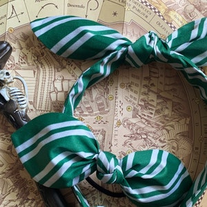 May include: A green and white striped fabric headband with a bow. The headband features a repeating pattern of green and white stripes. A decorative cane with a skeleton is visible in the background.