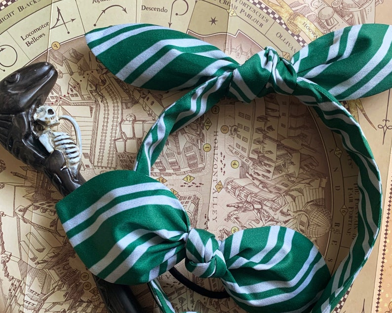 May include: A green and white striped fabric headband with a bow. The headband features a repeating pattern of green and white stripes. A decorative cane with a skeleton is visible in the background.