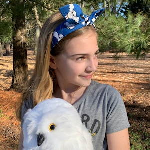 May include: A young person wearing a blue headband with a white owl pattern and a grey shirt with the word "Bird" on it. They are holding a white stuffed owl with yellow eyes.