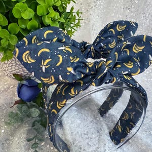 May include: A navy blue headband and bow with a yellow banana pattern. The bow is tied on top of the headband. The background is a sparkly white surface with a green plant and a blue flower.