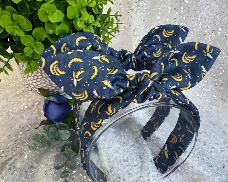 May include: A navy blue headband and bow with a yellow banana pattern. The bow is tied on top of the headband. The background is a sparkly white surface with a green plant and a blue flower.