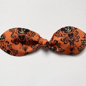 K&ouml;nnte beinhalten: Orangefarbenes Stoffhaarband mit einem schwarz-wei&szlig;en Muster der Haunted Mansion. Das Muster zeigt das ikonische Herrenhaus, Geister und die Worte "Haunted Mansion".