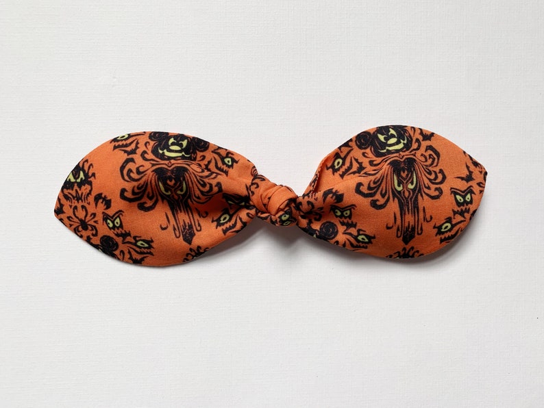 K&ouml;nnte beinhalten: Orangefarbenes Stoffhaarband mit einem schwarz-wei&szlig;en Muster der Haunted Mansion. Das Muster zeigt das ikonische Herrenhaus, Geister und die Worte "Haunted Mansion".
