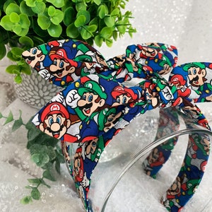 May include: A blue headband with a bow featuring a colorful pattern of Mario and Luigi characters. The headband is made of fabric and has a clear plastic band. The bow is tied at the top of the headband.