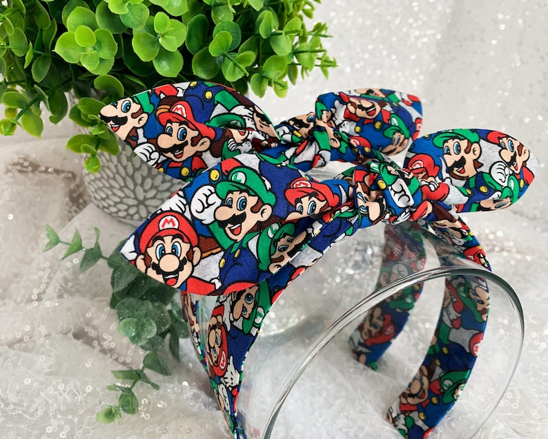 May include: A blue headband with a bow featuring a colorful pattern of Mario and Luigi characters. The headband is made of fabric and has a clear plastic band. The bow is tied at the top of the headband.