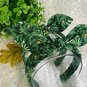 Pu&ograve; includere: Fascia per capelli in tessuto verde e giallo con un nodo al centro. Il tessuto ha un motivo di foglie verdi e occhi gialli.