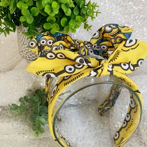 May include: Yellow headband with a Minion character print. The headband is tied in a bow and is made of fabric.