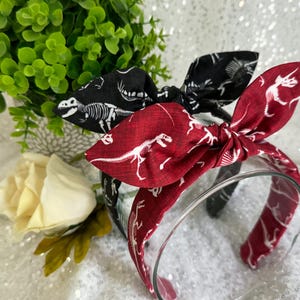 May include: Two fabric headbands with dinosaur skeletons printed on them. One headband is black with white skeletons and the other is red with white skeletons.