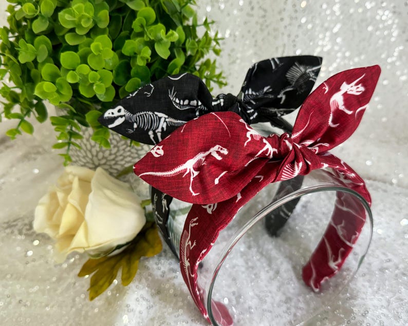 May include: Two fabric headbands with dinosaur skeletons printed on them. One headband is black with white skeletons and the other is red with white skeletons.