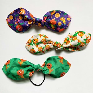 May include: Three fabric hair bows with a cartoon character and fruit design. The bows are purple, white, and green.