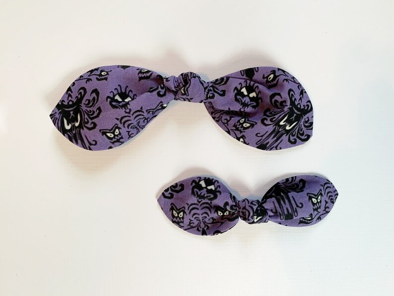 K&ouml;nnte beinhalten: Zwei lilafarbene Haarb&auml;nder mit einem schwarzen und wei&szlig;en Muster eines Spukhauses. Das gr&ouml;&szlig;ere Band ist zu einem Knoten gebunden, w&auml;hrend das kleinere Band zu einer Schleife gebunden ist.