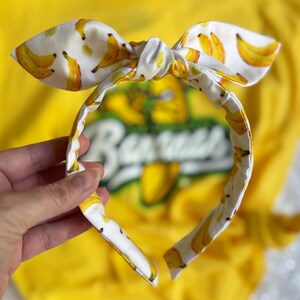 May include: A white headband with a bow, patterned with yellow bananas. The headband is held in front of a yellow shirt with the word "Bananas" visible. The bow is tied in the center.