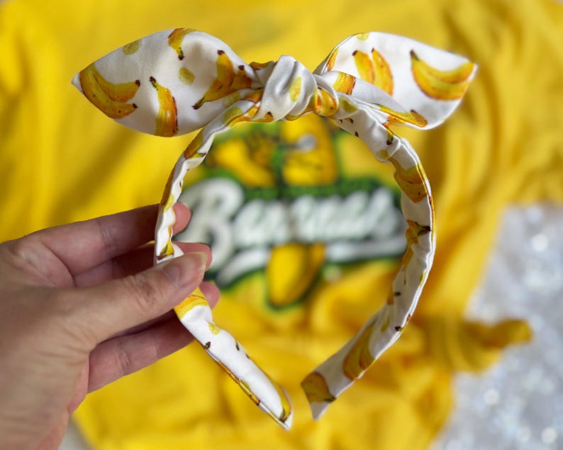 May include: A white headband with a bow, patterned with yellow bananas. The headband is held in front of a yellow shirt with the word "Bananas" visible. The bow is tied in the center.