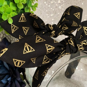 May include: Black headband with a gold patterned design of the Deathly Hallows symbol from the Harry Potter series.