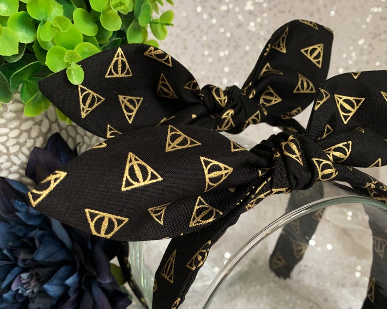 May include: Black headband with a gold patterned design of the Deathly Hallows symbol from the Harry Potter series.