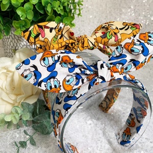 May include: A white headband with a blue and white Donald Duck print on one side and a yellow and brown Goofy print on the other side.