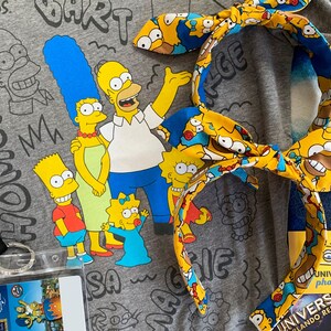 May include: A gray t-shirt featuring The Simpsons family with a matching headband. The headband has a yellow background with Homer Simpson's face. A Universal Studios Orlando Resort photo card is also visible.