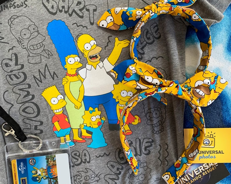 May include: A gray t-shirt featuring The Simpsons family with a matching headband. The headband has a yellow background with Homer Simpson's face. A Universal Studios Orlando Resort photo card is also visible.