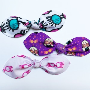 May include: Three purple and white fabric hair bows with different patterns. One bow has a pattern of eyes with glasses, another has a pattern of a girl with glasses and a wand, and the last bow has a pattern of glasses.