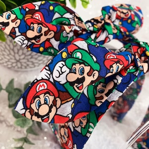 May include: A blue headband with a bow featuring a repeating pattern of Mario and Luigi characters. The characters are in red, green, and white on a blue background. The headband is a fun accessory for fans of the video game.