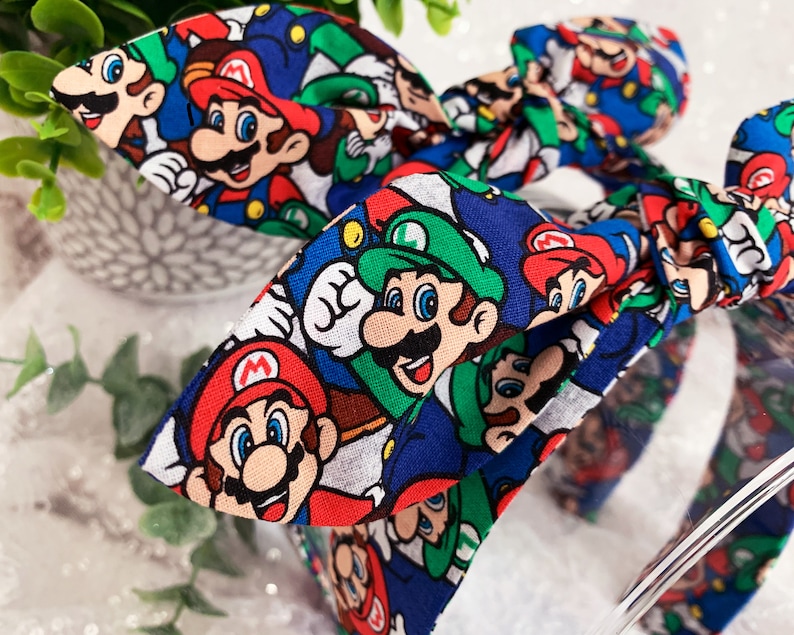 May include: A blue headband with a bow featuring a repeating pattern of Mario and Luigi characters. The characters are in red, green, and white on a blue background. The headband is a fun accessory for fans of the video game.