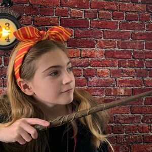 May include: A young person wearing a red and yellow striped headband with a bow, holding a brown and gold wand with a glowing tip. The wand is pointed towards the right side of the image. A sign with the text "PLATFORM 9 3/4" is visible in the background.