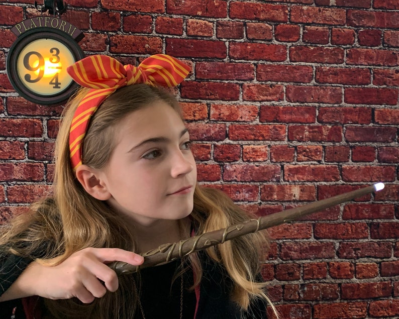 May include: A young person wearing a red and yellow striped headband with a bow, holding a brown and gold wand with a glowing tip. The wand is pointed towards the right side of the image. A sign with the text "PLATFORM 9 3/4" is visible in the background.