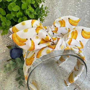 May include: A white headband with a large bow featuring a repeating pattern of yellow bananas. The bananas have brown and yellow details. The headband is made of fabric and has a clear plastic band.