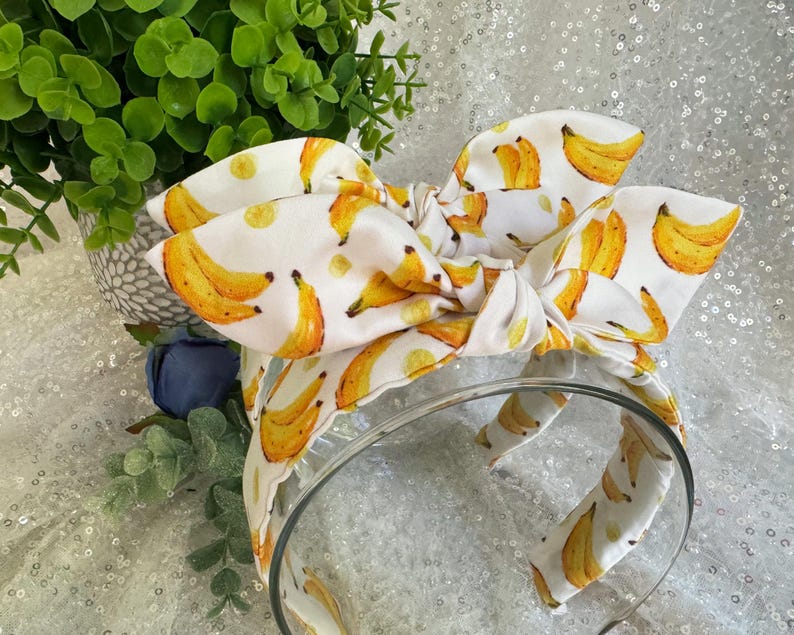May include: A white headband with a large bow featuring a repeating pattern of yellow bananas. The bananas have brown and yellow details. The headband is made of fabric and has a clear plastic band.
