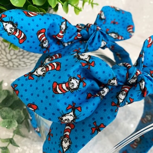May include: A blue headband with a white polka dot pattern and a repeating image of the Cat in the Hat. The headband is tied in a bow.