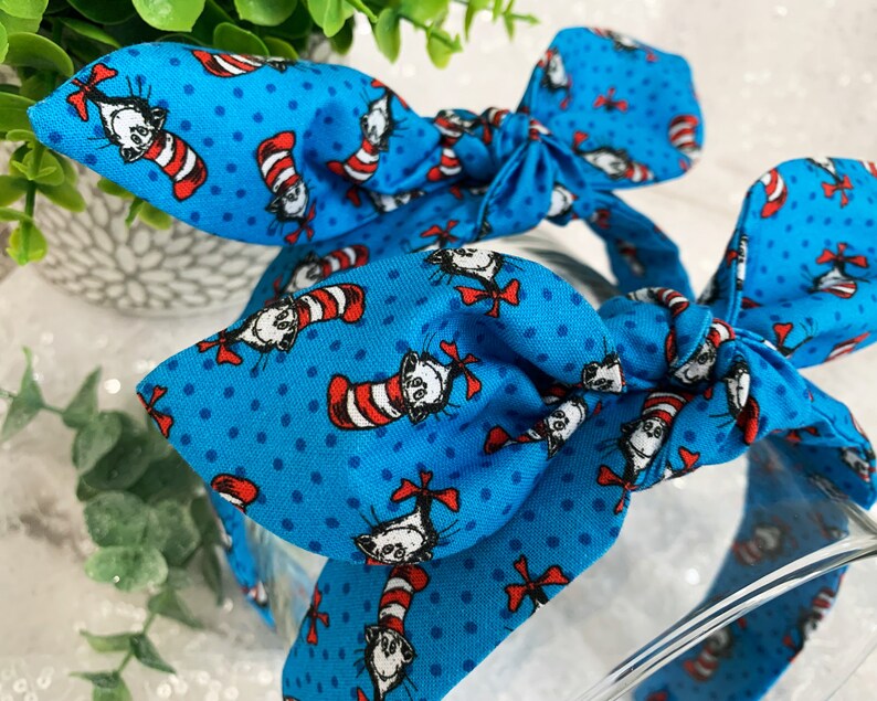 May include: A blue headband with a white polka dot pattern and a repeating image of the Cat in the Hat. The headband is tied in a bow.