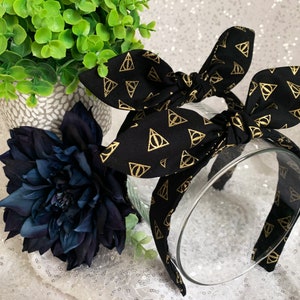 May include: Black headband with a gold patterned fabric featuring the Deathly Hallows symbol. The headband is tied into a bow.