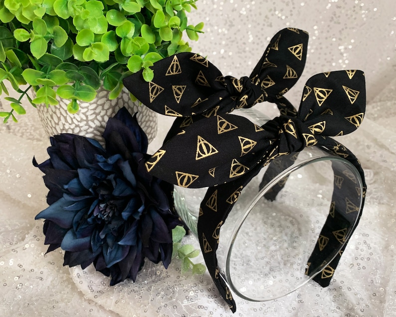 May include: Black headband with a gold patterned fabric featuring the Deathly Hallows symbol. The headband is tied into a bow.