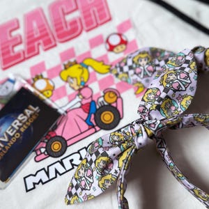 May include: White t-shirt with a pink and white graphic of Princess Peach in a kart, the word "PEACH" above, and "MARIO" below. A light purple headband with a Princess Peach pattern and a Universal Orlando Resort pass are also visible.