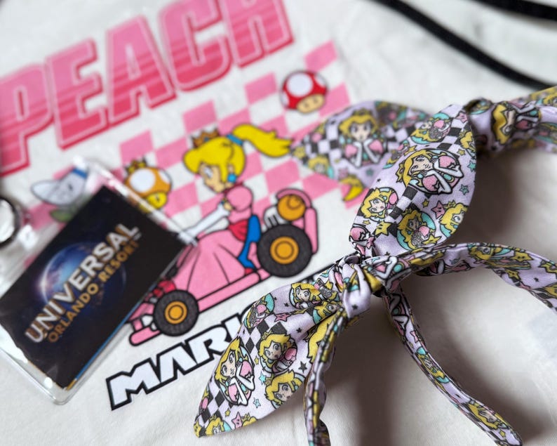 May include: White t-shirt with a pink and white graphic of Princess Peach in a kart, the word "PEACH" above, and "MARIO" below. A light purple headband with a Princess Peach pattern and a Universal Orlando Resort pass are also visible.