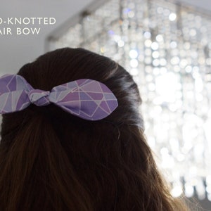 K&ouml;nnte beinhalten: Eine violette und blaue geometrische gemusterte Haarschleife, die zu einem Knoten gebunden ist. Die Schleife ist in den Haaren einer Person gebunden.