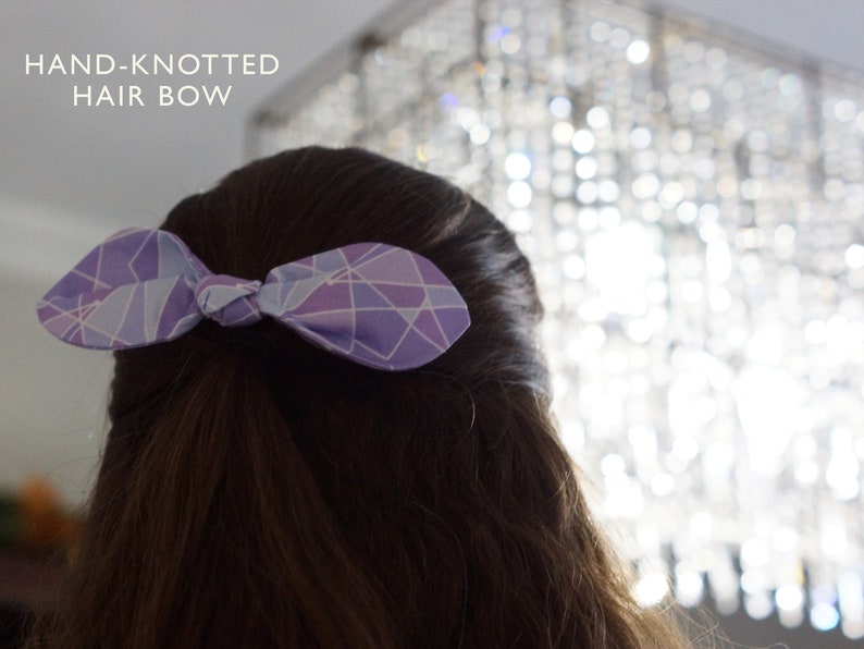 K&ouml;nnte beinhalten: Eine violette und blaue geometrische gemusterte Haarschleife, die zu einem Knoten gebunden ist. Die Schleife ist in den Haaren einer Person gebunden.