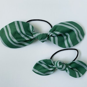 May include: Two green and white striped fabric hair ties. The larger hair tie is tied in a bow and the smaller hair tie is tied in a smaller bow.