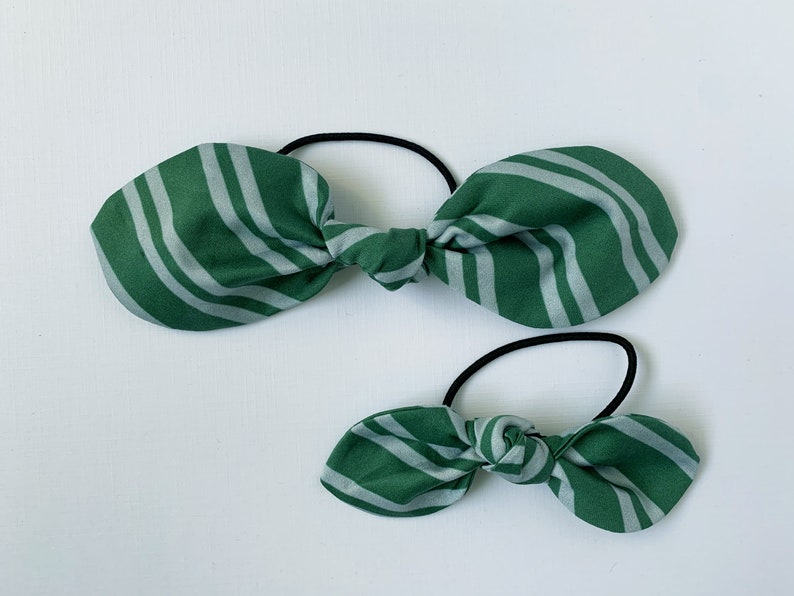 May include: Two green and white striped fabric hair ties. The larger hair tie is tied in a bow and the smaller hair tie is tied in a smaller bow.